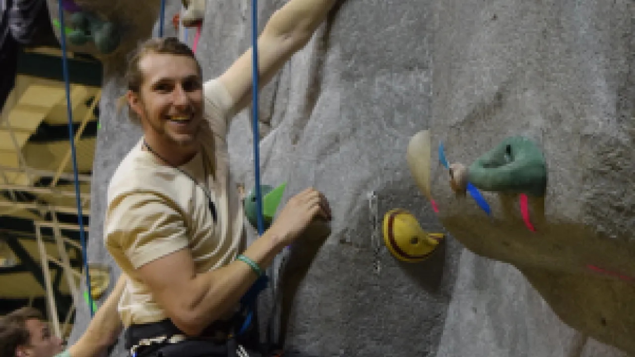 Male-presenting person hangs from a rope attached to his harness as he climbs the indoor climbing wall. 