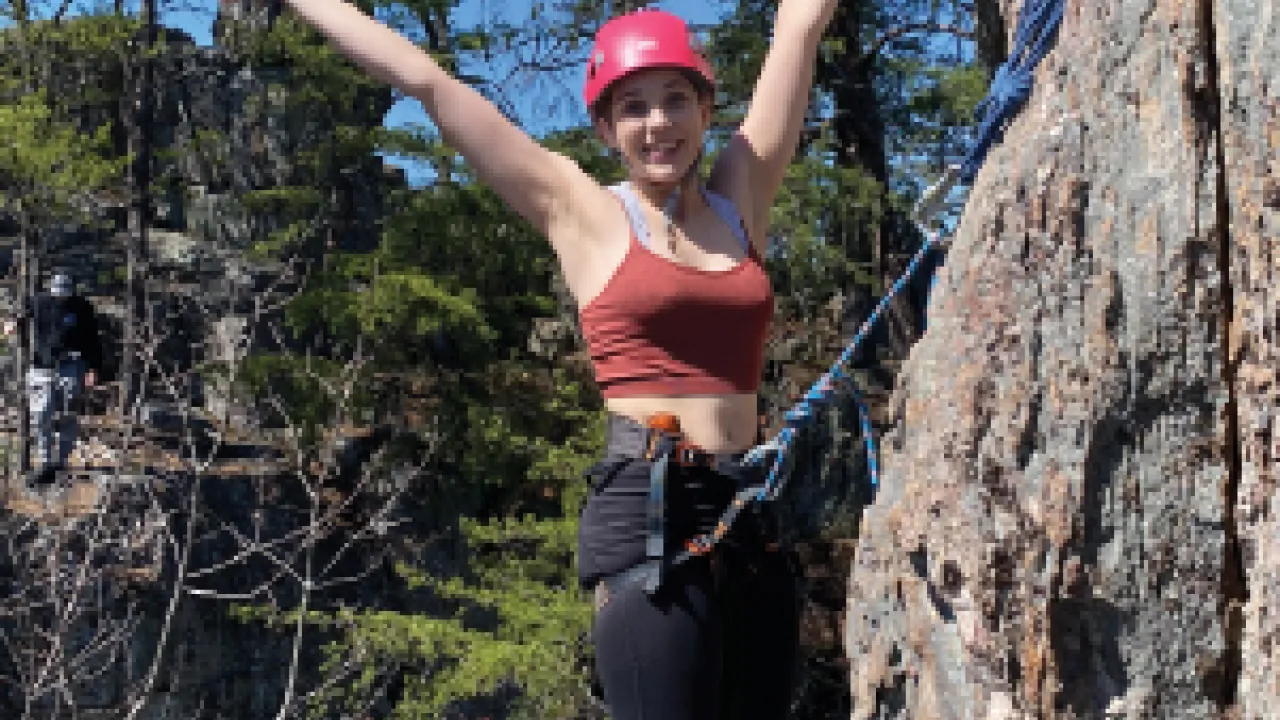 Femme person with arms spread wide, smiling at the camera while attached by rope to her harness to the rock face.