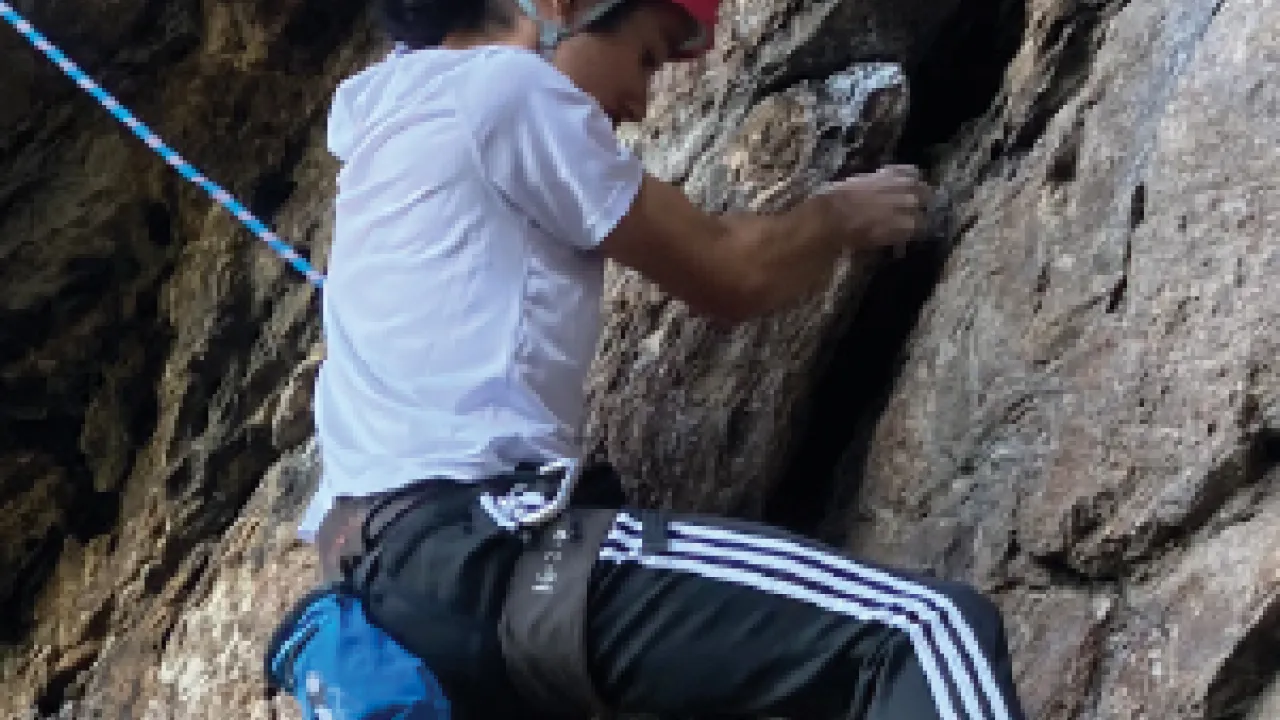 Person with long hair tied in a low bun, wearing a pick climbing helmet, looks down at their feet while climbing a cliff face.
