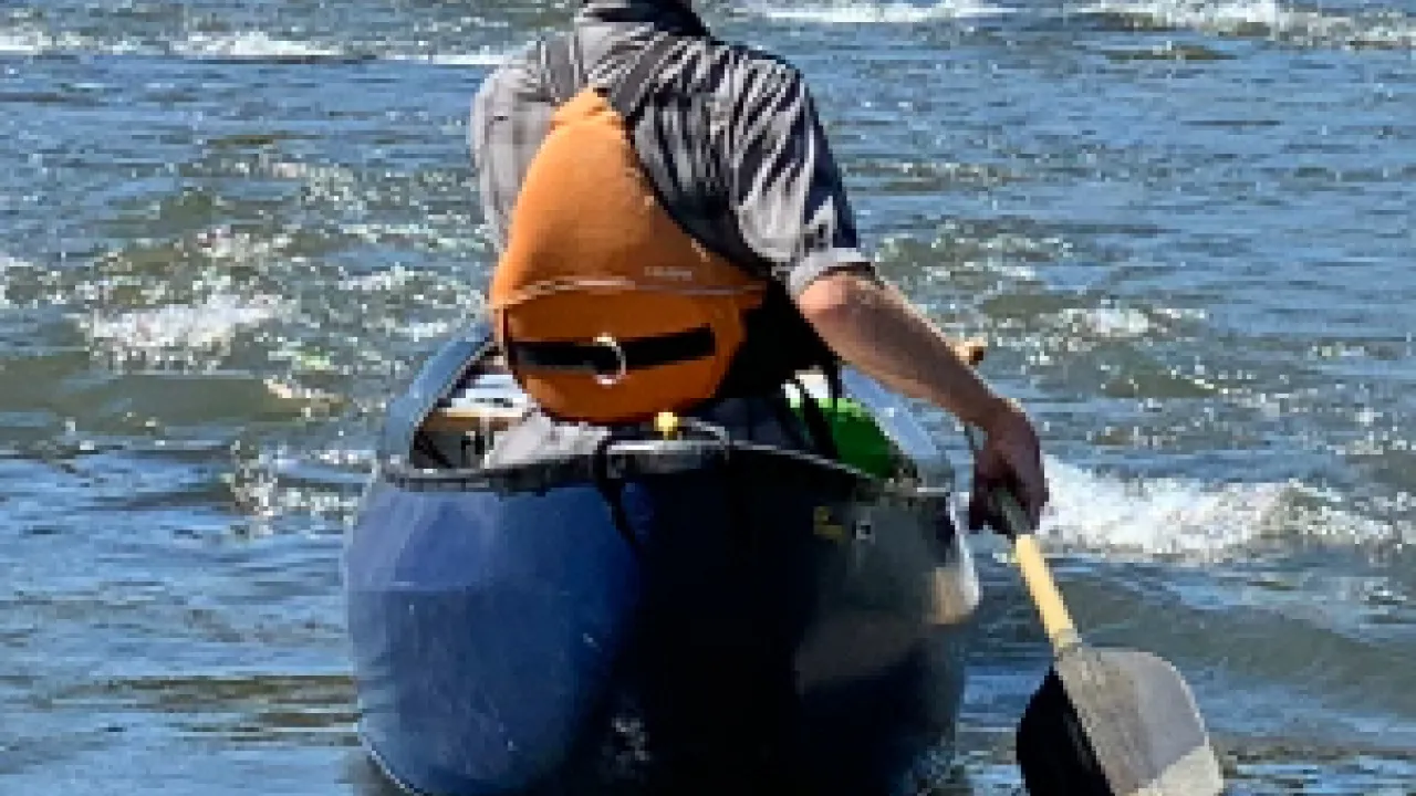 Person with back to the camera, paddles a canoe on a waterway, wearing bright yellow helmet and orange PFD.