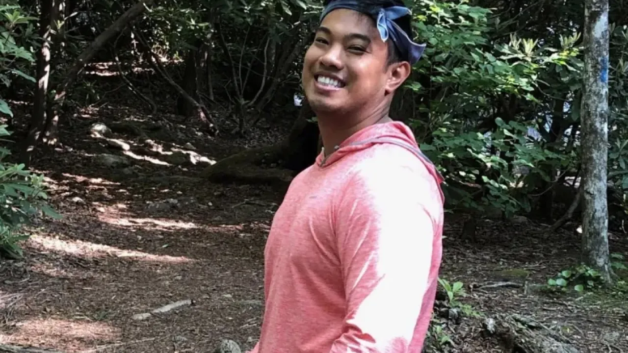 male-presenting person smiling at the camera while standing against a backdrop of green trees.