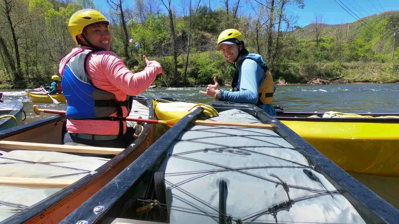 Male-presenting person smiles at camera with their thumb up while sitting in a whitewater canoe
