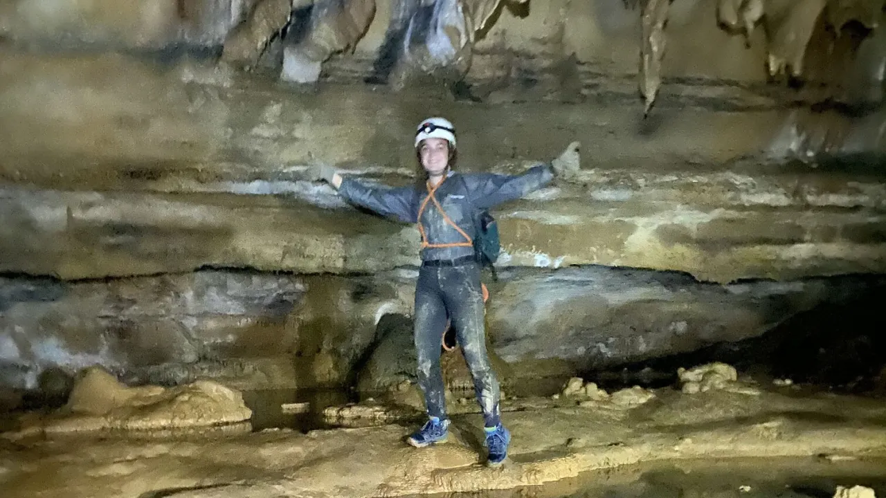 Femme person with arms spread wide, wearing a blue jump suit, white helmet, and headlamp. She is standing inside a cave with stalactites overhead and an underground river at her feet.