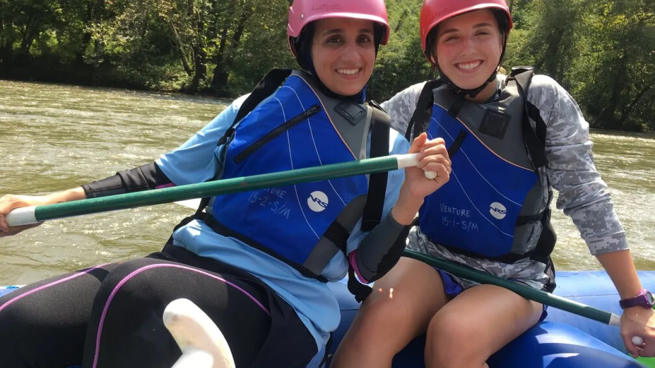 Two people in a raft smiling, both wearing blue lifejackets with a river and greenery behind them.