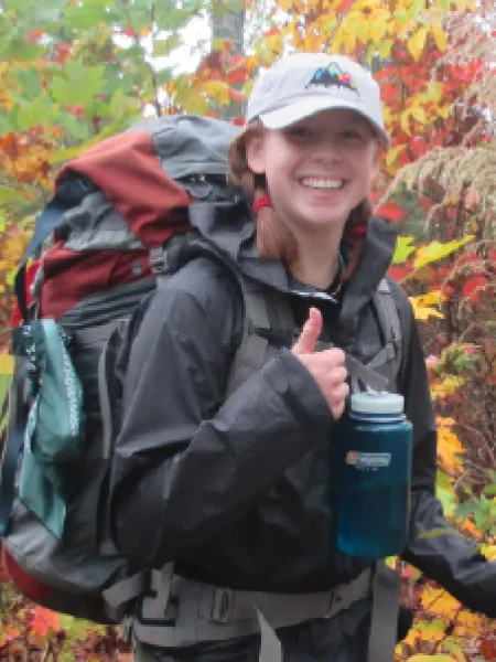 Femme person wearing a ball cap and backpack, giving a thumbs up to the camera with a big smile.