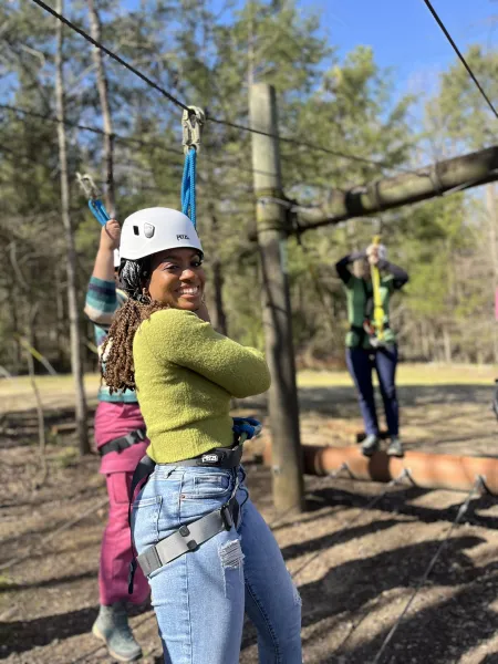 Female presenting person smiles at the camera while wearing safety equipment at a ropes course