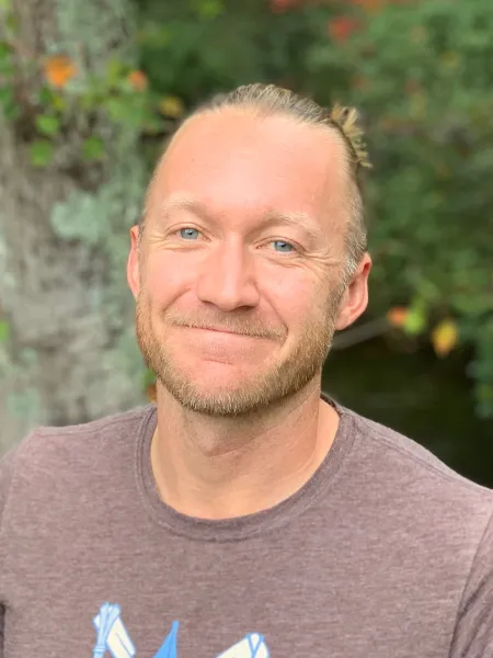 Smiling, male-presenting person in front of a green trees