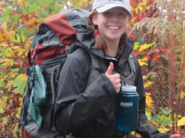 Femme person wearing a ball cap and backpack, giving a thumbs up to the camera with a big smile.