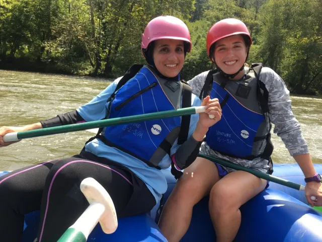 Two people in a raft smiling, both wearing blue lifejackets with a river and greenery behind them.