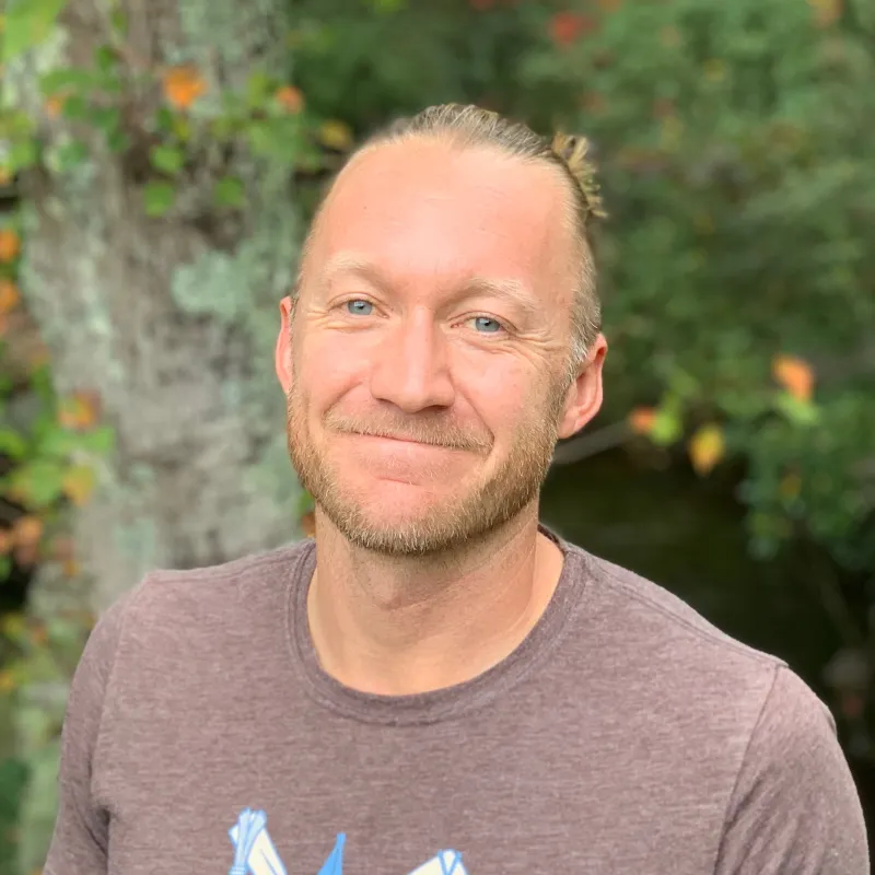 Smiling, male-presenting person in front of a green trees